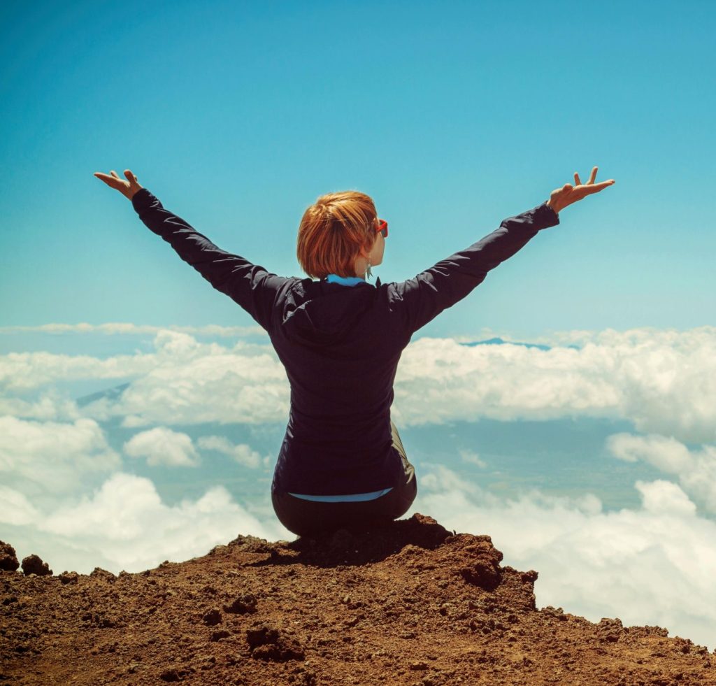 A woman enjoys a scenic view atop a cliff in Kula, Hawaii, surrounded by clouds and blue sky.